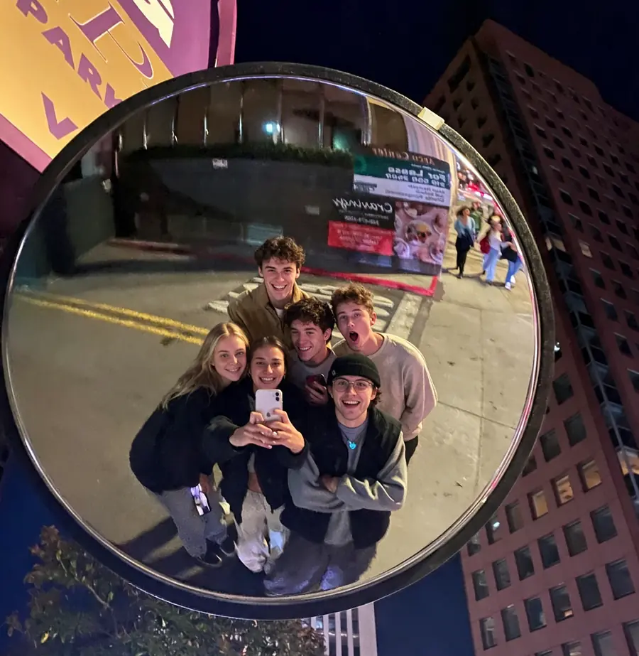 A group of friends at night taking a selfie in a round street mirror, buildings all around