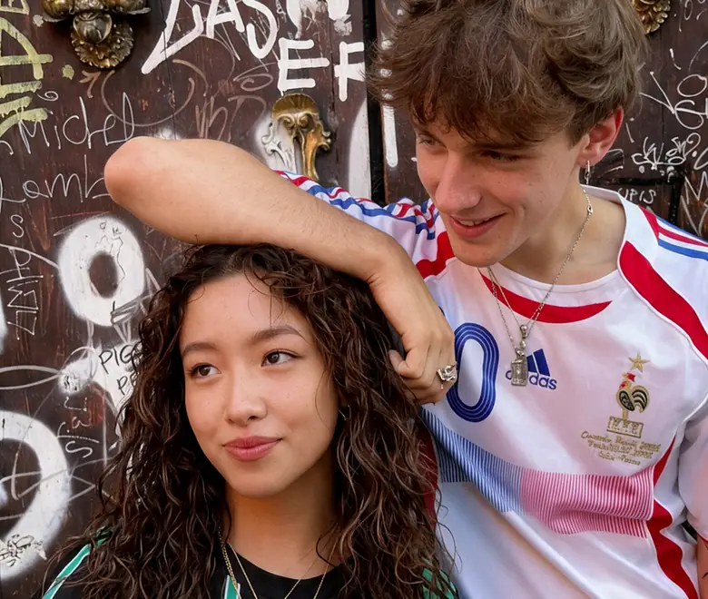 Un chico apoya el brazo en la cabeza de una chica frente a una puerta llena de grafitis; los dos sonríen